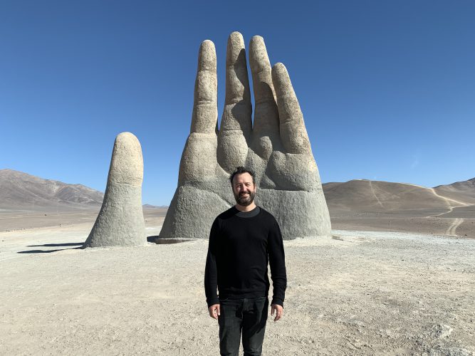 Man standing in front of a statue of a hand coming out of the ground that is twice as tall as he is